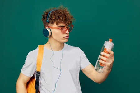 a funny man stands on a gray background in pink glasses, with a bright backpack on his back and examines a water bottle for sportsの写真素材