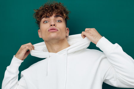 a handsome man stands on a green background in a white hoodie and poses relaxed, adjusting the hood on his head with his handsの写真素材