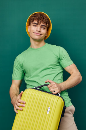 a cute, happy man stands on a green background, dressed in a yellow panama hat, holding a bright travel suitcase in his hand, looking pleasantly into the camera Travel and vacation themeの写真素材