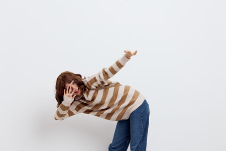 a funny, emotional woman in a stylish striped sweater and blue jeans leans to the side, pointing her index finger upwards towards the empty space, and with the other hand covers her faceの写真素材