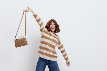 a beautiful, happy, joyful woman in stylish clothes stands on a light background and holds up her beige bag in a joyful gestureの写真素材