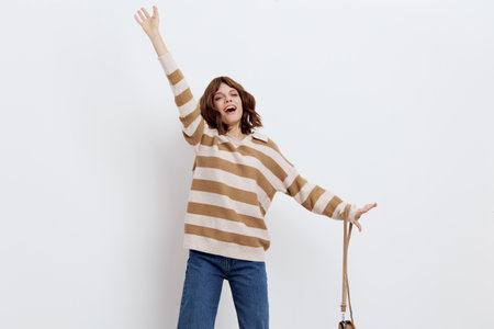 joyful woman is jumping up and down merrily and energetically, posing against a white background in a striped sweater and blue jeans with a beige bag on her shoulder, joyfully raising her hands up.の写真素材