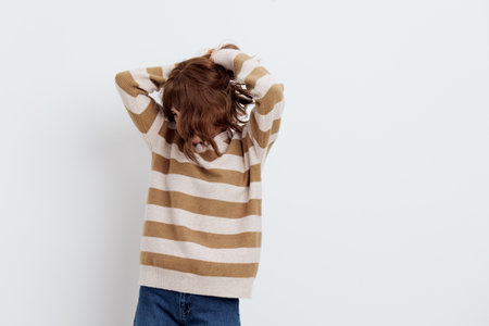 a beautiful, pleasant woman stands in a stylish striped sweater on a light background and her hair covers her face, she corrects them with her handsの写真素材