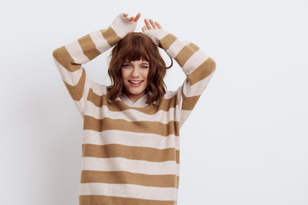 a sweet, happy, funny woman stands on a white background in a stylish striped sweater and holds her hands behind her head, smiling pleasantly and making a funny face looking at the cameraの写真素材