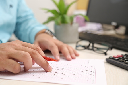 Man is hand on the filled OMR sheet with pen. Picture of computer, flower pot and glasses.