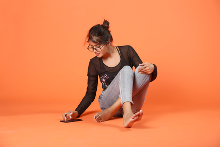 Beautiful girl is sitting on the floor is using phone. Isolated on orange background.の写真素材
