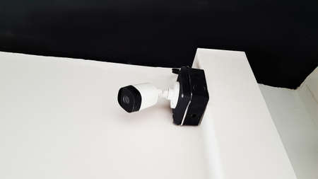 A security camera installed at the top of the white pole and wall with the dark ceiling.の写真素材