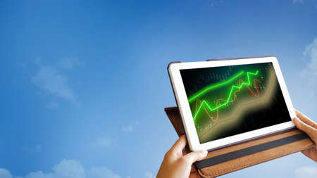 Tablet with stock market trading graph on screen, holding in hands on blue background, concept of using technology device to trade on stock exchange appsの写真素材