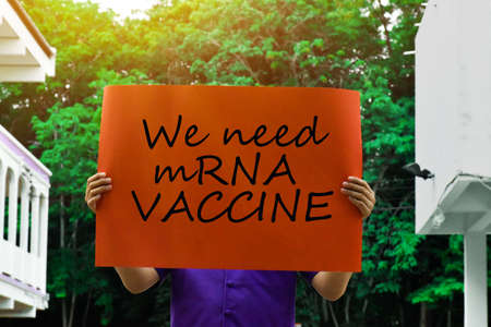 Orange label with text "We need mRNA VACCINE" holding in hands with green trees and the sunlight in backgroundの写真素材