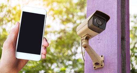 A hand holding a mobile phone is placed on a picture of a CCTV camera at a home. This highlights the integration of modern technology in home security.の写真素材