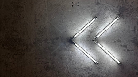 An arrangement of fluorescent light tubes forming an arrow pointing to the left, a gradient of gray to dark tones, offering a sleek and modern backdrop copy space for text.の写真素材