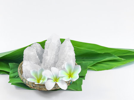 Clear and pristine alum cubes resting in a bamboo basket alongside leaves, set against a white background, showcasing the natural purity and traditional presentation of the substance.の写真素材