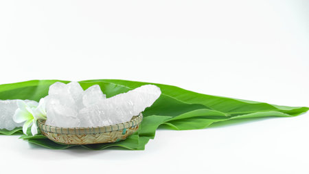 Clear and pristine alum cubes resting in a bamboo basket alongside leaves, set against a white background, showcasing the natural purity and traditional presentation of the substance.の写真素材