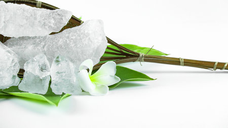 Clear and pristine alum cubes resting in a bamboo basket alongside leaves, set against a white background, showcasing the natural purity and traditional presentation of the substance.の写真素材