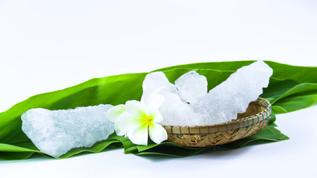 Clear and pristine alum cubes resting in a bamboo basket alongside leaves, set against a white background, showcasing the natural purity and traditional presentation of the substance.の写真素材