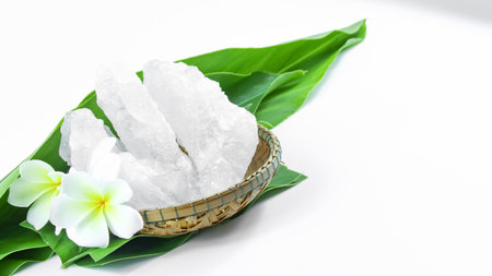 Clear and pristine alum cubes resting in a bamboo basket alongside leaves, set against a white background, showcasing the natural purity and traditional presentation of the substance.の写真素材