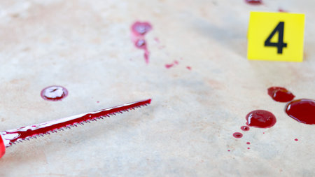 A bloodstained stainless steel handsaw lying on a concrete floor, with a blurred yellow marker number 4 and scattered blood drops, symbolizing a violent crime scene under investigation.の写真素材