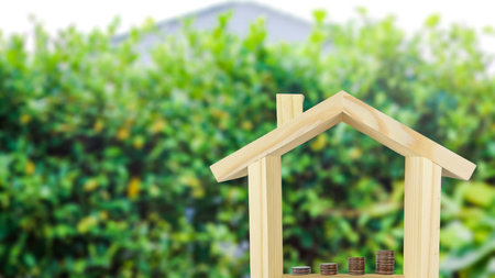 A wooden house model with coins arranged in ascending order inside, set against a blurred background of bushes, symbolizing financial growth and investment for home ownership.の写真素材