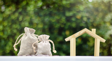 Brown burlap sacks filled with coins and some scattered on a white surface and wooden house model placed nearby, with blurred background of bushes, growth in savings and financial planning.の写真素材