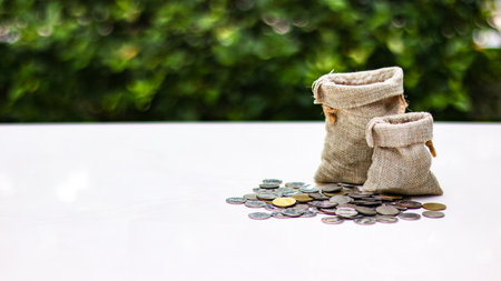 A burlap sack standing and another lying with coins spilling out onto a white surface, set against a blurred background of bushes, representing financial planning and budgeting.の写真素材