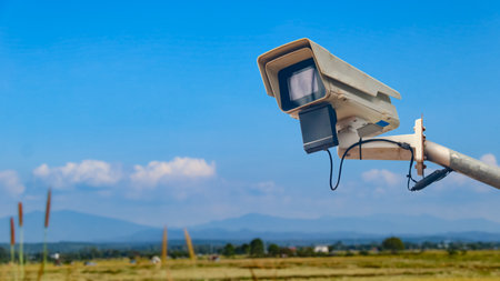 Old CCTV mounted on steel railing with blurred rice fields and mountains, symbolizing security,...