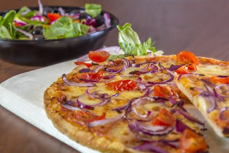 Pizza with tomatoes and onions on a brown wooden table with salad in the backgroundの写真素材