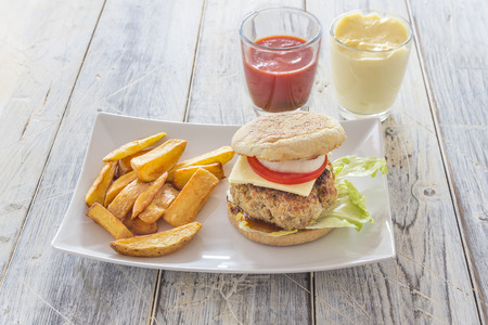 homemade burger and french fries on a white plate with ketchup and mayonnaise in glasses on a wooden tableの写真素材