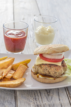 homemade burger and french fries on a white plate with ketchup and mayonnaise in glasses on a wooden tableの写真素材