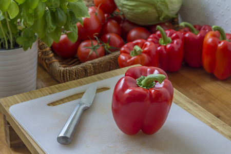 Red Pepper and a knife on a board in a kitchen with more red pepper and other vegetables in the backgroundの写真素材