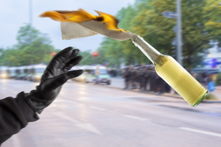 Arm in black sleeve with black leather glove thwowing a Molotov Cocktail with burning fuse, with police forces in the blurred backgroundの写真素材