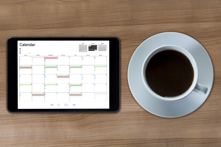 A tablet computer with a calendar displayed and a cup of coffee on a wooden desktopの写真素材