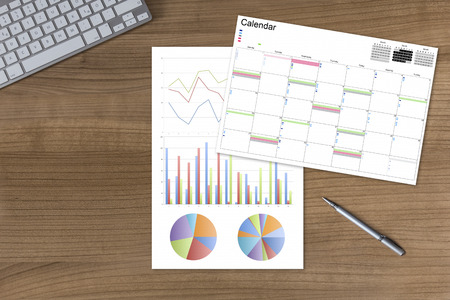 Calendar and a sheet with colorful diagrams on a wooden table with modern keyboard and silver penの写真素材