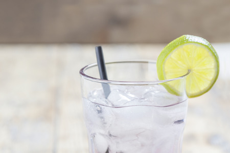 A glass filled with a transparent liquid, ice cubes and a lime slice with a straw on wooden tableの写真素材