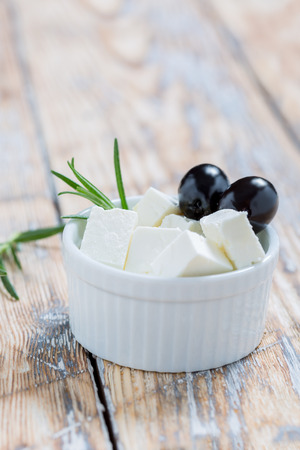 Feta slices with olives and rosemary in a white bowl on a rustic wooden tableの写真素材