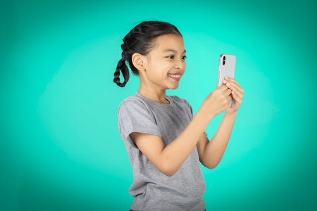 A little Asian girl in her right hand holds a mobile phone. and taking selfie with a mobile phone isolated on Green background. look smartphone front camera, photographing, record video message post online.の写真素材