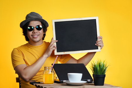 Young Man Holding Empty Blackboard While Working on Summer Vacation Season Against Yellow Background         の写真素材