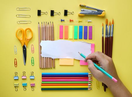Hand Holding Pencil on Torn Paper. School Stationary Flat Lay on Yellow Backgroundの写真素材