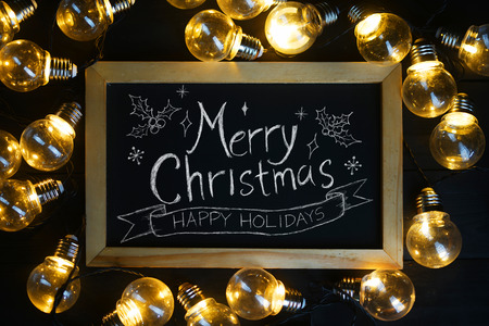Merry Christmas Typography on Blackboard Between Light Bulbs on Black Woodの写真素材