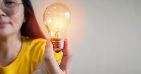 An Asian woman hand holding light bulb creative new ideas technology concept, Female smile happy looking light orange flare effect connection cream color background space for text.の写真素材