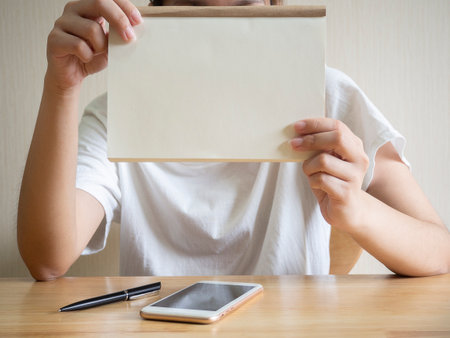 Woman sitting and holding white blank paper sheet mockup on wooden beige background. Checklist tablet plan concept.の写真素材