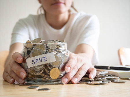 Close up of woman holding money jar with coins on a light brown wooden table. Investment business, retirement, finance and saving money for future concept.の写真素材