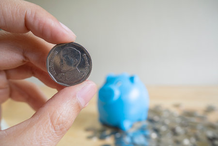 Close up of hand holding a coin with solf blur behide of blue piggy bank and standing on the pile of coins. Saving money concept.の写真素材