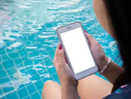 Young woman traveler relaxing and using a mobile phone by a hotel pool while traveling for summer vacation, isolated screen.の写真素材