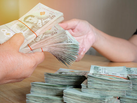 Handing over stacks of Thai banknote to other hand with soft light background. Money and finance concept.の写真素材