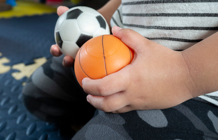 Asian boy sitting on foam carpet in living room and in his hands holding soccer balls and basketball, toys in the house.の写真素材
