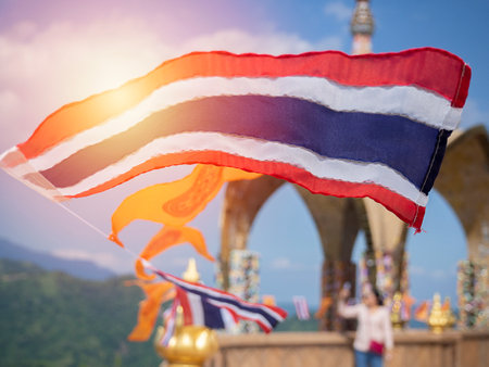 The Thailand national flag flies in front of a traditional Buddhist temple building and tourist is standing in the behide. Blue sky and mountain background.の写真素材