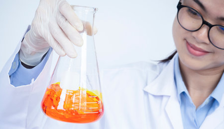 Erlenmeyer flask filled with orange liquid chemical held and examined by young female researcher scientist in laboratoryの写真素材