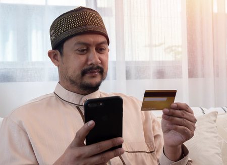 Internet Mobile Shopping Concept. Positive Asian Muslim man using cell phone holding looking at credit card purchasing things online sitting on sofa buying gifts in web store, full body lengthの写真素材