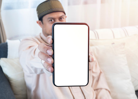 Young Asian muslim man in traditional clothes sitting at sofa in the living room and showing mobile phone with isolated screen.の写真素材