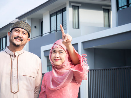 Happy homeowners Asian muslim couple pointing their new house. First property, smiling and enjoy relocation day at outdoor. Bank loan, mortgage for young family concept.の写真素材